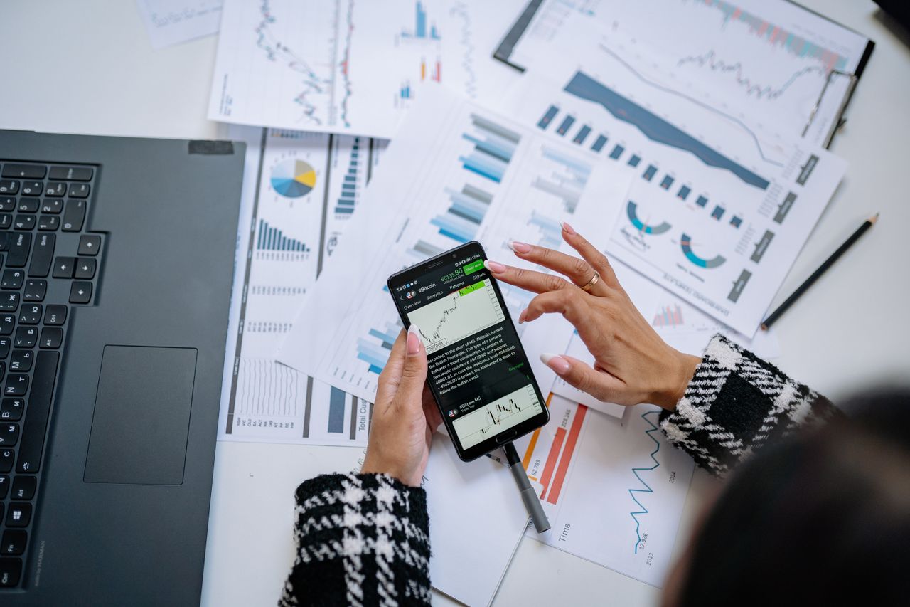 Une femme d’affaires examine des graphiques financiers sur un smartphone, avec un ordinateur portable et des documents.